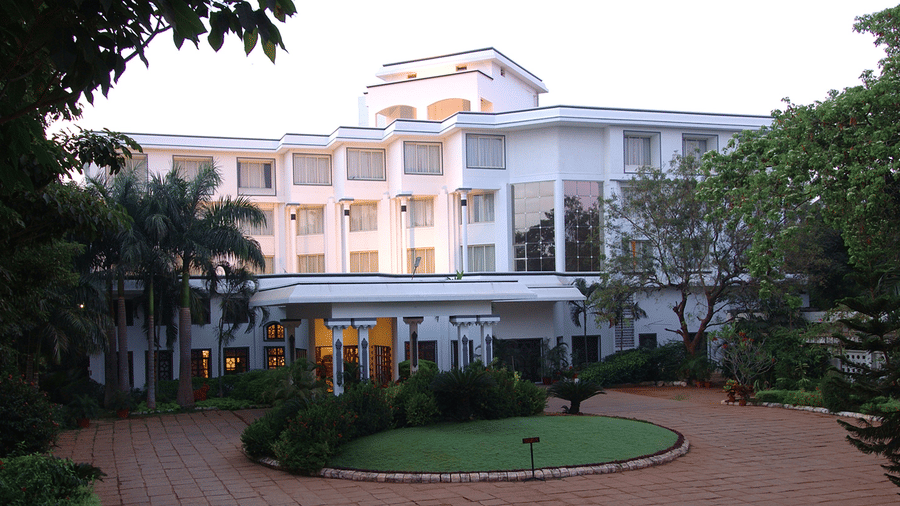 Facade view of Sangam Hotel, Thanjavur, with a pathway, a manicured garden and trees in view - Thanjavur Hotel Contact