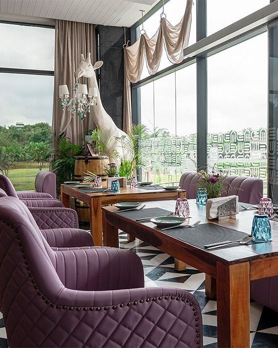 Indoor restaurant seating at Karma Chalets with purple chairs, wooden tables, and large glass windows overlooking greenery.