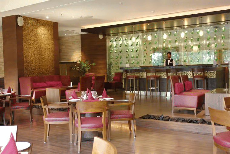 Restaurant dining area at Retreat with wooden chairs and pink upholstered seating arranged around tables with a backlit bar counter and decorative wall panels visible in the background.