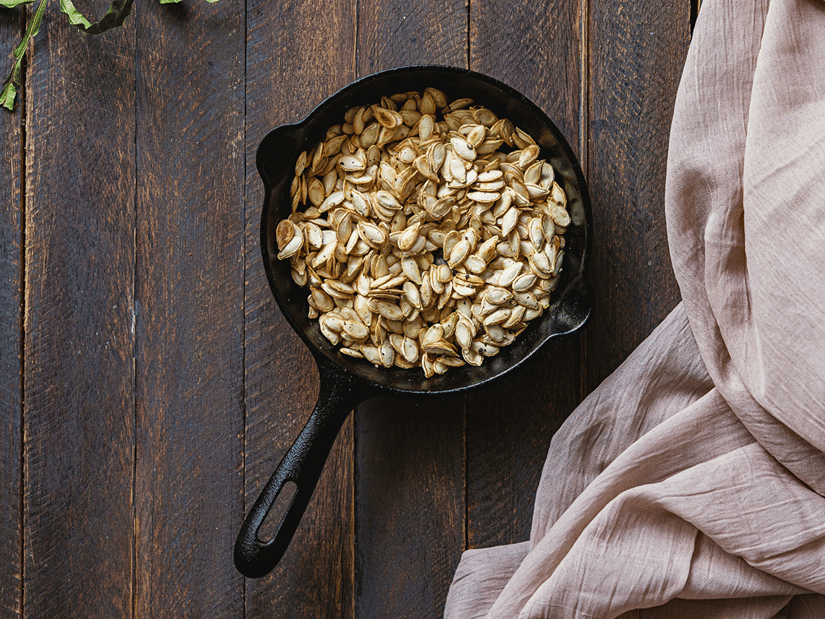 Roasted pumpkin seeds in a cast-iron skillet on a dark wooden table beside a pale pink linen cloth