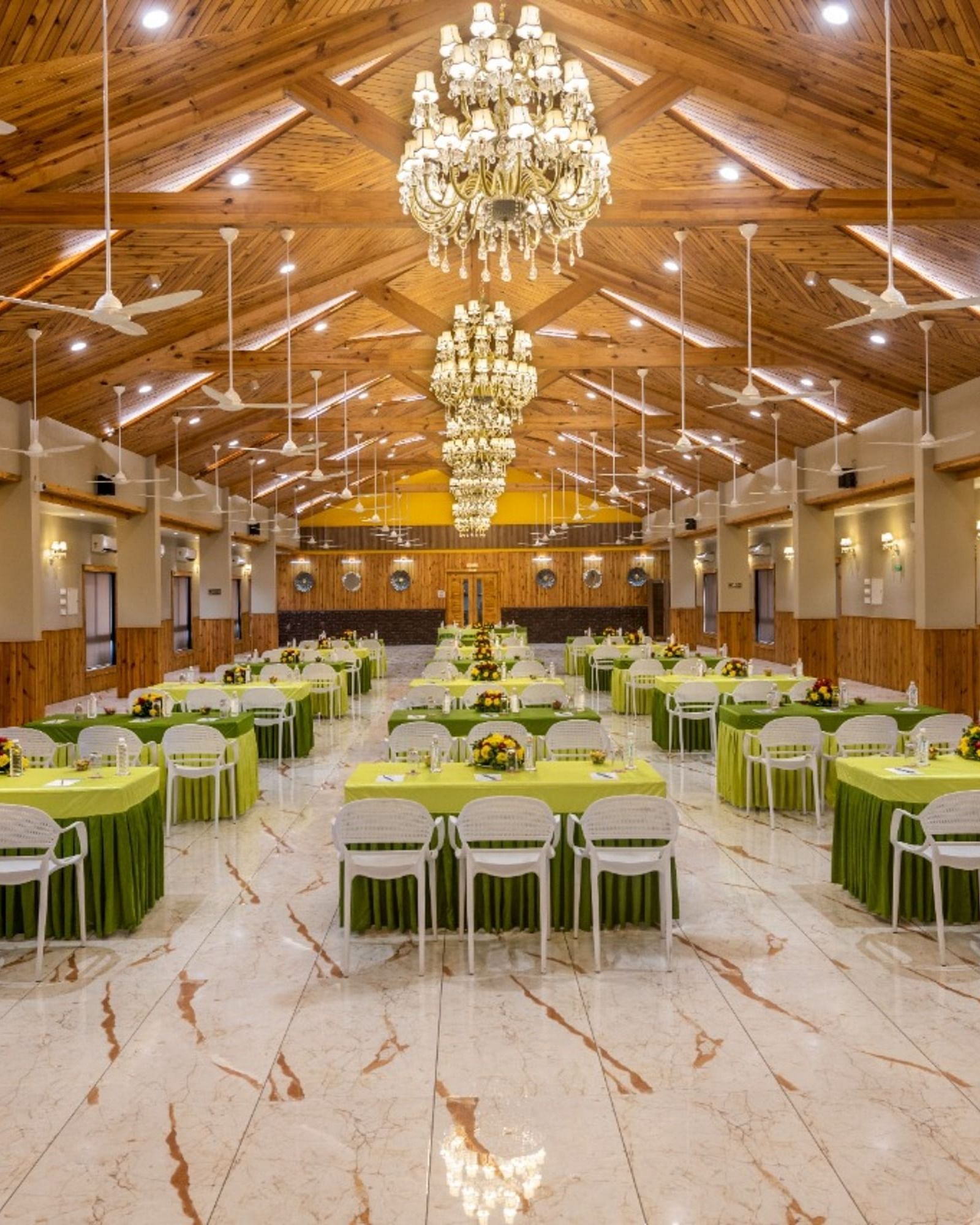 A well-lit banquet hall with multiple tables and chairs set for an event at Angel Lords Eco Inn Amreli.