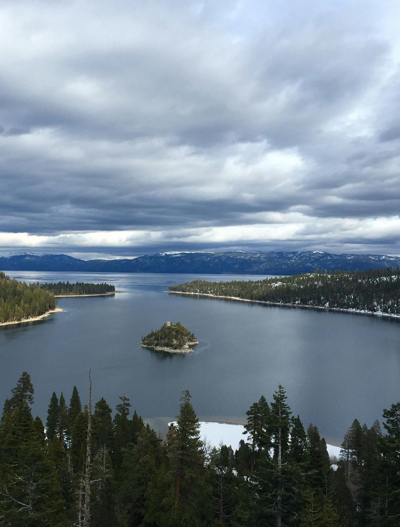 View of a lake with pine trees in the foreground on a cloudy day.