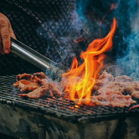 An action shot of meat grilling over an open flame with visible smoke and a person's hand holding tongs.