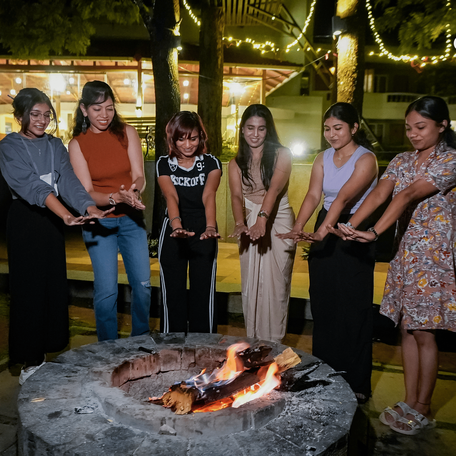 People standing by campfire at night at Grand Palace, Yercaud, with logs, firepit, outdoor setting, vegetation, lit faces and group reflection on experience.