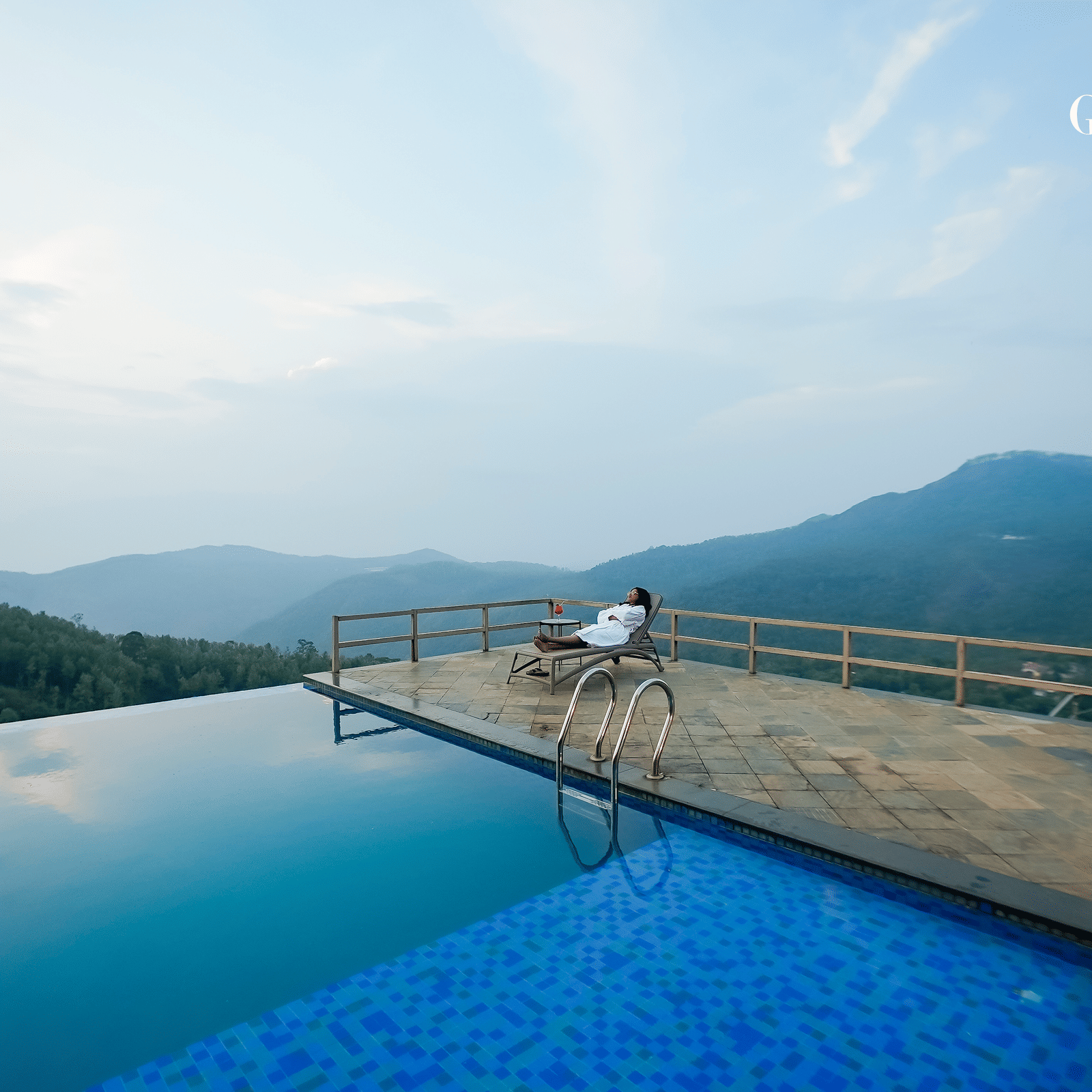 View of infinity pool at Grand Palace, Yercaud with deck, railing, distant hills, water surface, platform and wide sky above.