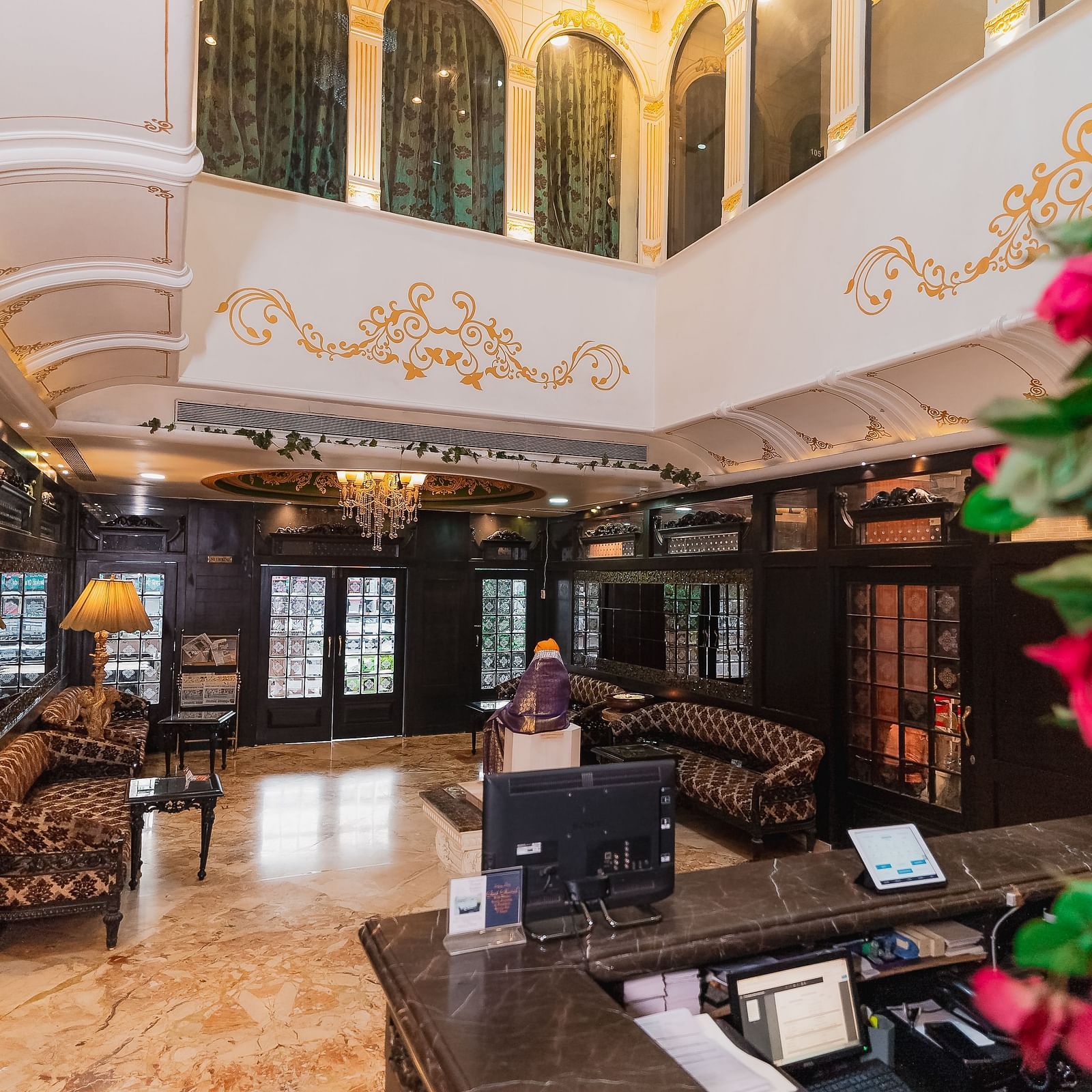 Hotel Ashok Nagpur featuring a wooden check-in counter with a computer and other equipment, a blurred figure in the background, and a floral arrangement on the side.