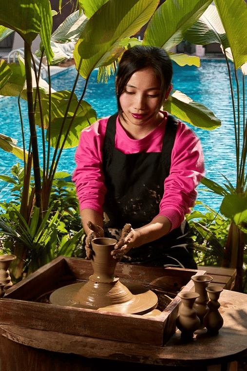 Woman in a pink top and black apron making pottery on a wheel outdoors, surrounded by lush plants, with a bright blue pool in the background.