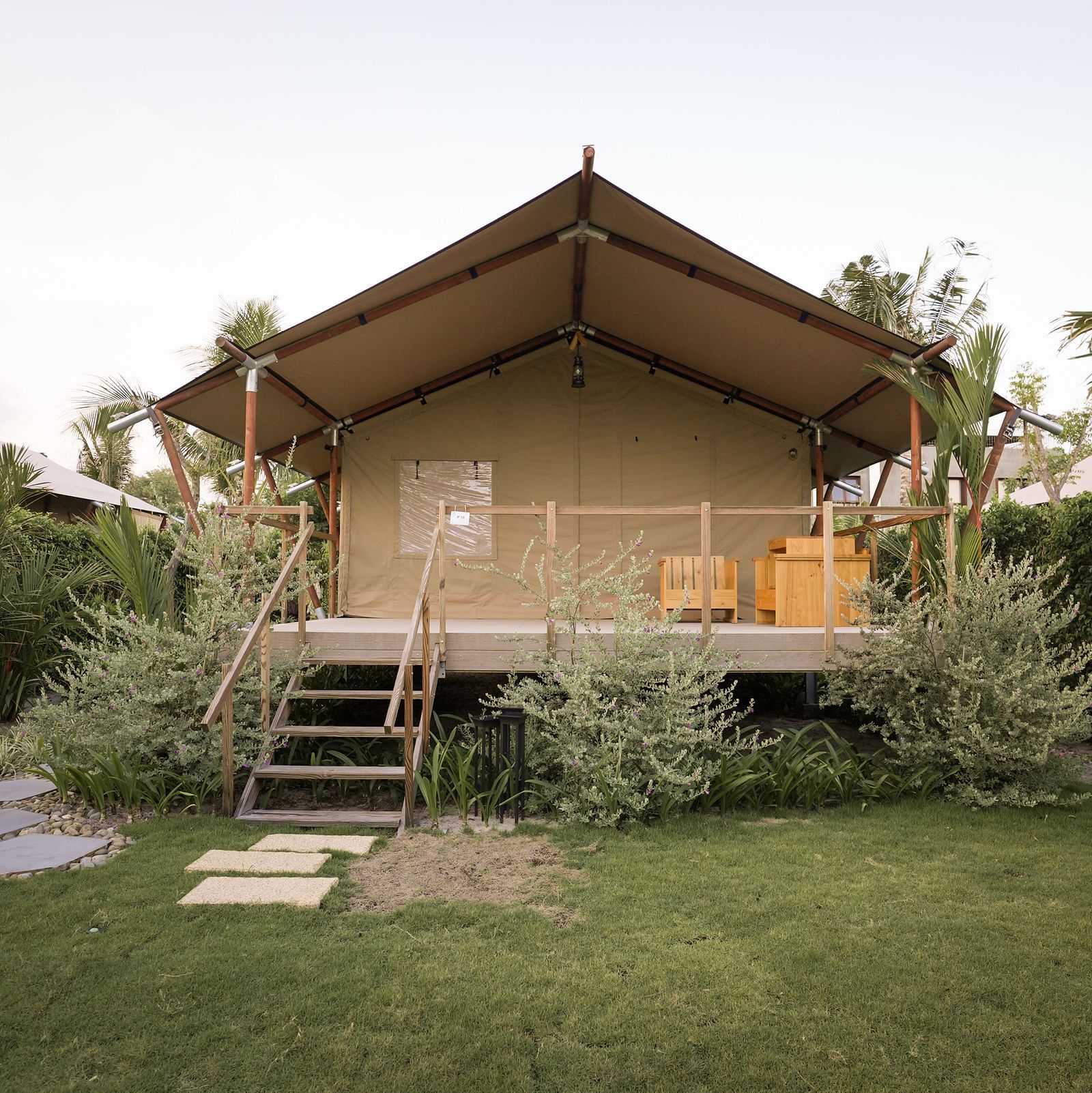 A wide lawn view of the Peaceful Tent at WoCAL Retreat Ke Ga, with stone steps leading to a charming canvas tent set against a backdrop of flowering shrubs and tall tropical trees.