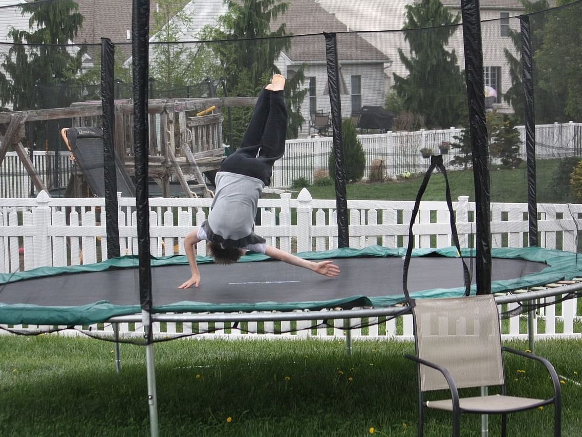 A kid jumping on a trampoline surrounded with standalone buildings and blue fencing.