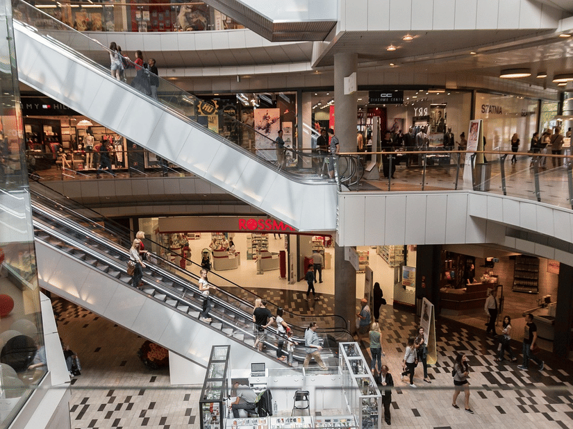 An interior view of a multi-level shopping mall featuring crisscrossing escalators with people riding them, surrounded by storefronts and walkways.