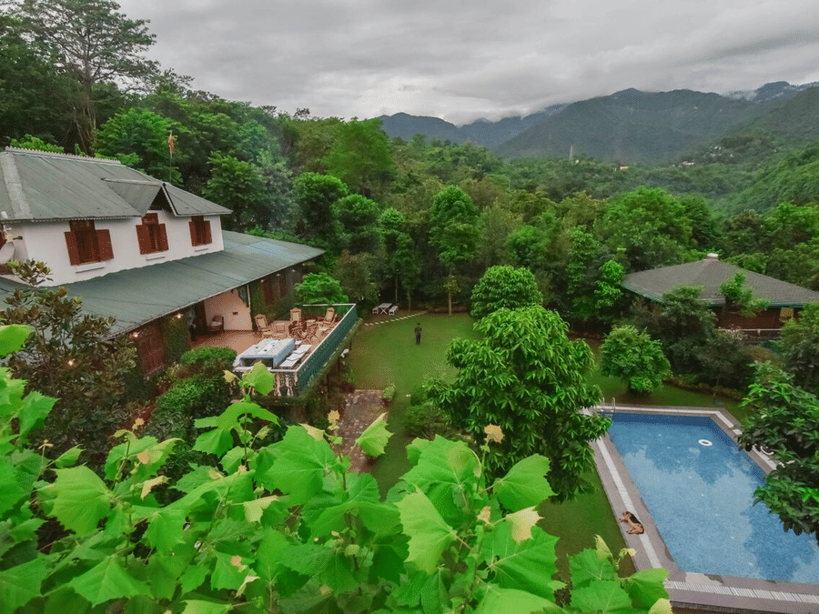 Facade of Shaheen Bagh - A Luxury Boutique Resort & Spa in Dehradun surrounded by trees with a swimming pool next to the lawn area and mountain ranges in the background.
