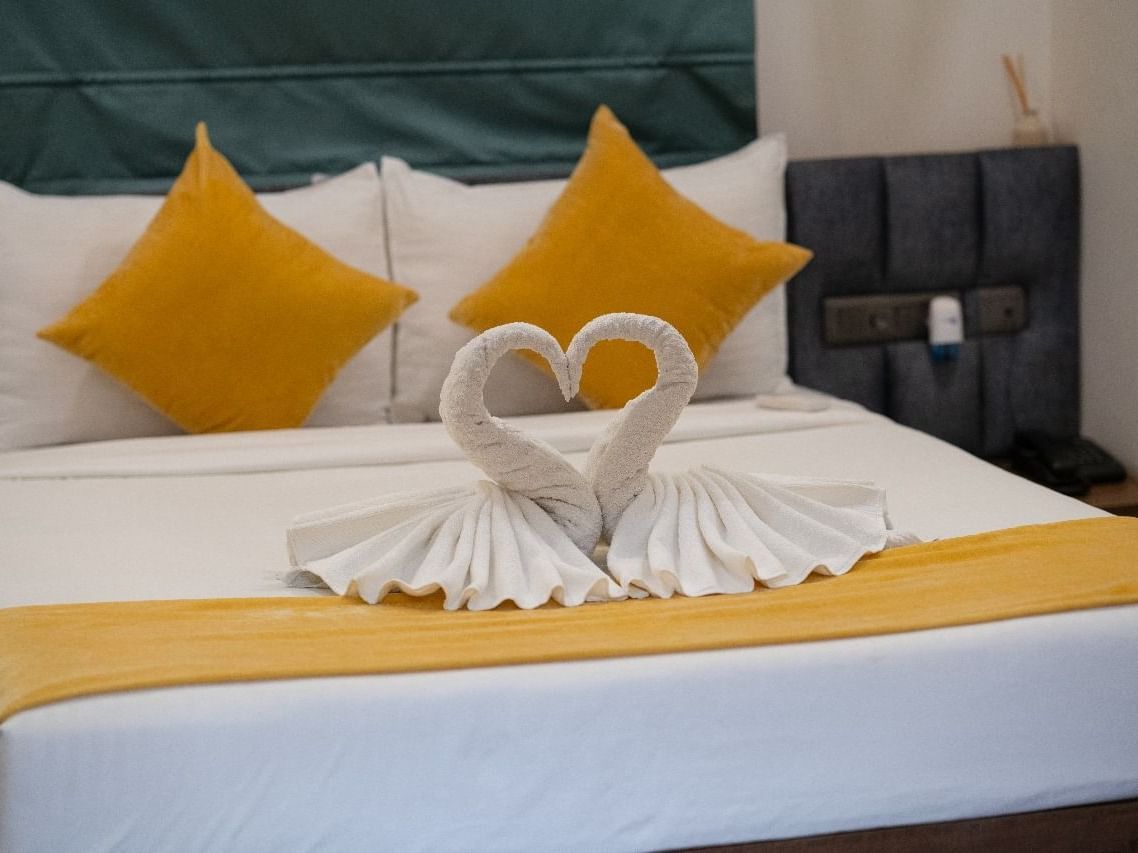 A room with a double bed, yellow pillows, and neatly arranged towels shaped into a heart, creating a romantic ambience at Hotel Kyzen Smartstays – Smart, Reliable, Kyzen