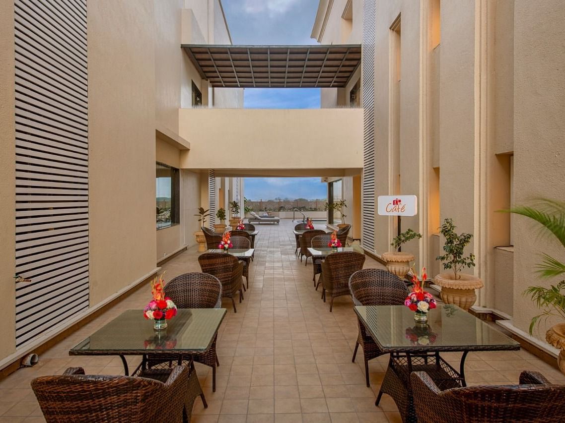 A well-lit outdoor seating area with multiple tables and chairs arranged along a pathway, featuring potted plants and a glimpse of the sky in the background, suggesting a relaxed dining space at Lords Inn Motikhavdi, Jamnagar.