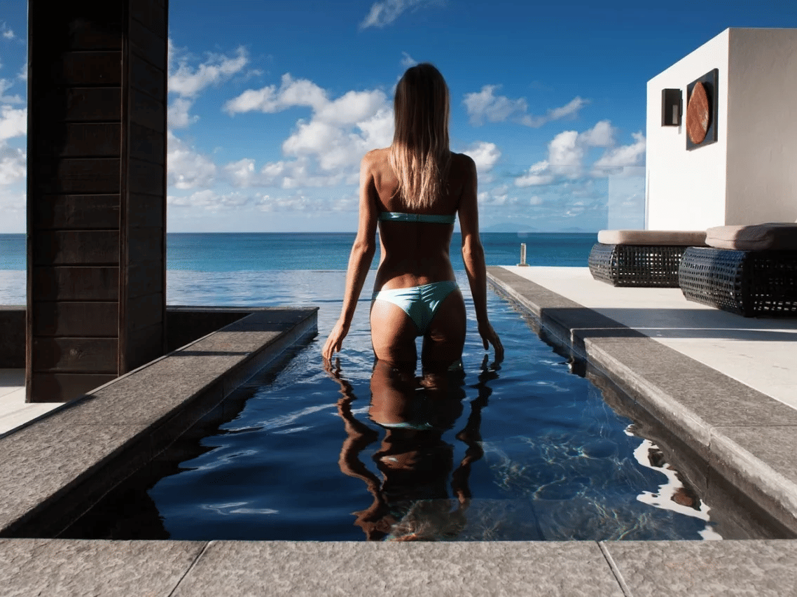 A woman in a light bikini stands in a narrow plunge pool, back to the camera, facing the vast blue ocean under a bright sky.