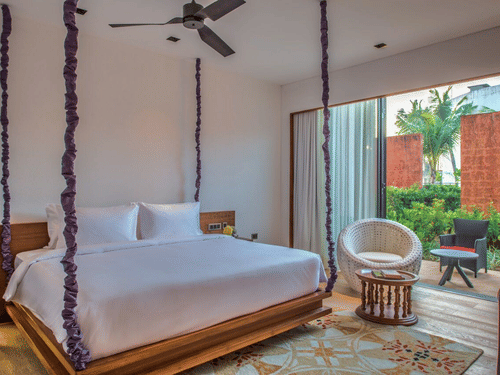 The Deluxe Patio King Room with a large wooden bed hanging from the ceiling on purple rope vines, and an armchair at Azaya Beach Resort, Goa.