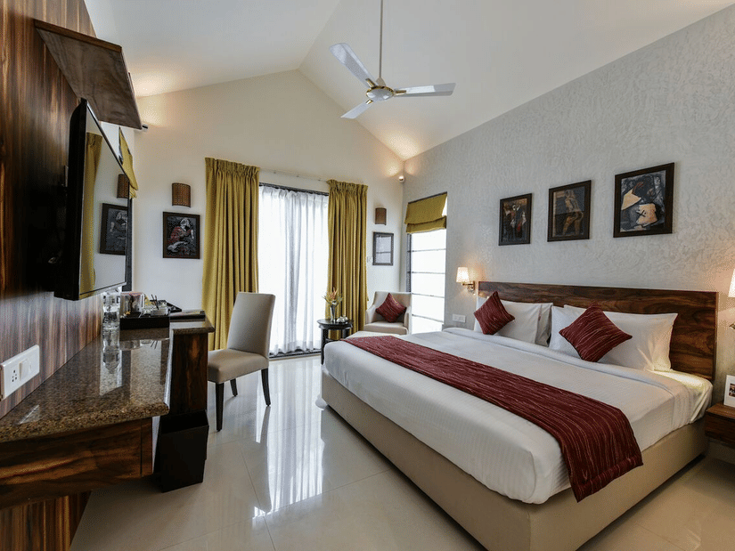 Family room at Tropicana Resort & Spa, Alibaug, featuring a king size bed with white linens and red accents. The room includes vaulted ceilings, a ceiling fan, a wooden headboard, a wall-mounted TV, and a desk area near a large window with gold curtains.