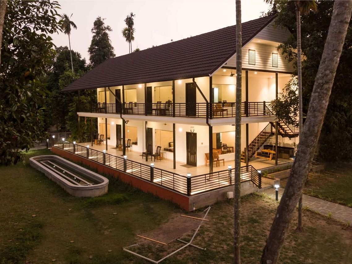 An exterior view of a well-lit, multi-story building at dusk, the Lhasa Ayurveda & Wellness Resort, with surrounding greenery.