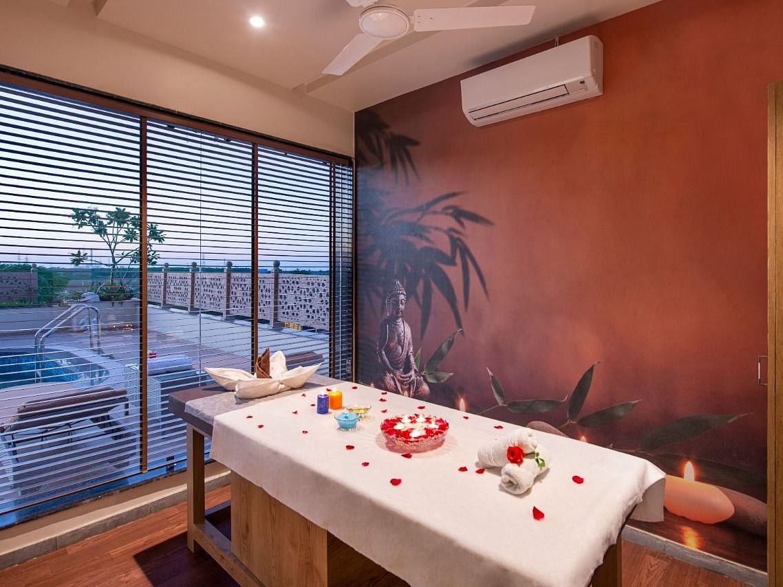 A serene spa treatment room featuring a massage table draped with a white sheet and adorned with red rose petals and decorative items, set against a warm-toned wall with a subtle mural and with natural light coming through large windows overlooking an outd