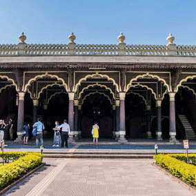 Front view of the Tippu Sultan's Palace in Bengaluru, during a sunny day with a pathway lined with green and yellow-hued bushes.