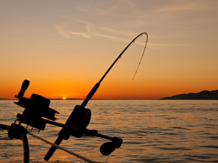 A fishing rod positioned over water at sunset with the sky turning orange.