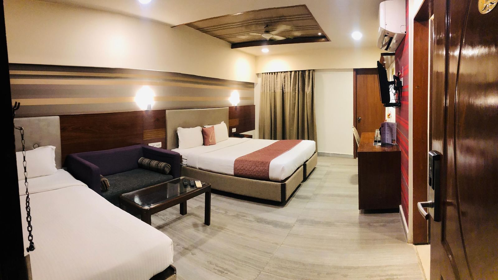 Hotel room interior showcasing a double bed, a dark-colored sofa, a glass-topped coffee table, and a work desk with a chair  | Astra Hotels and Suites Kadubeesanahalli
