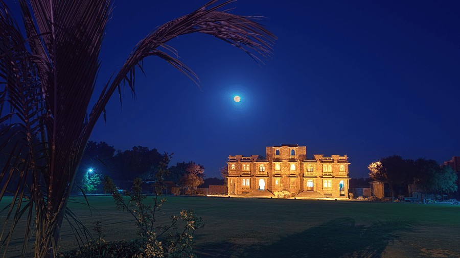 A multi-storey stone building at Garh Govind stands illuminated on a lawn under a dark night sky featuring a bright moon.