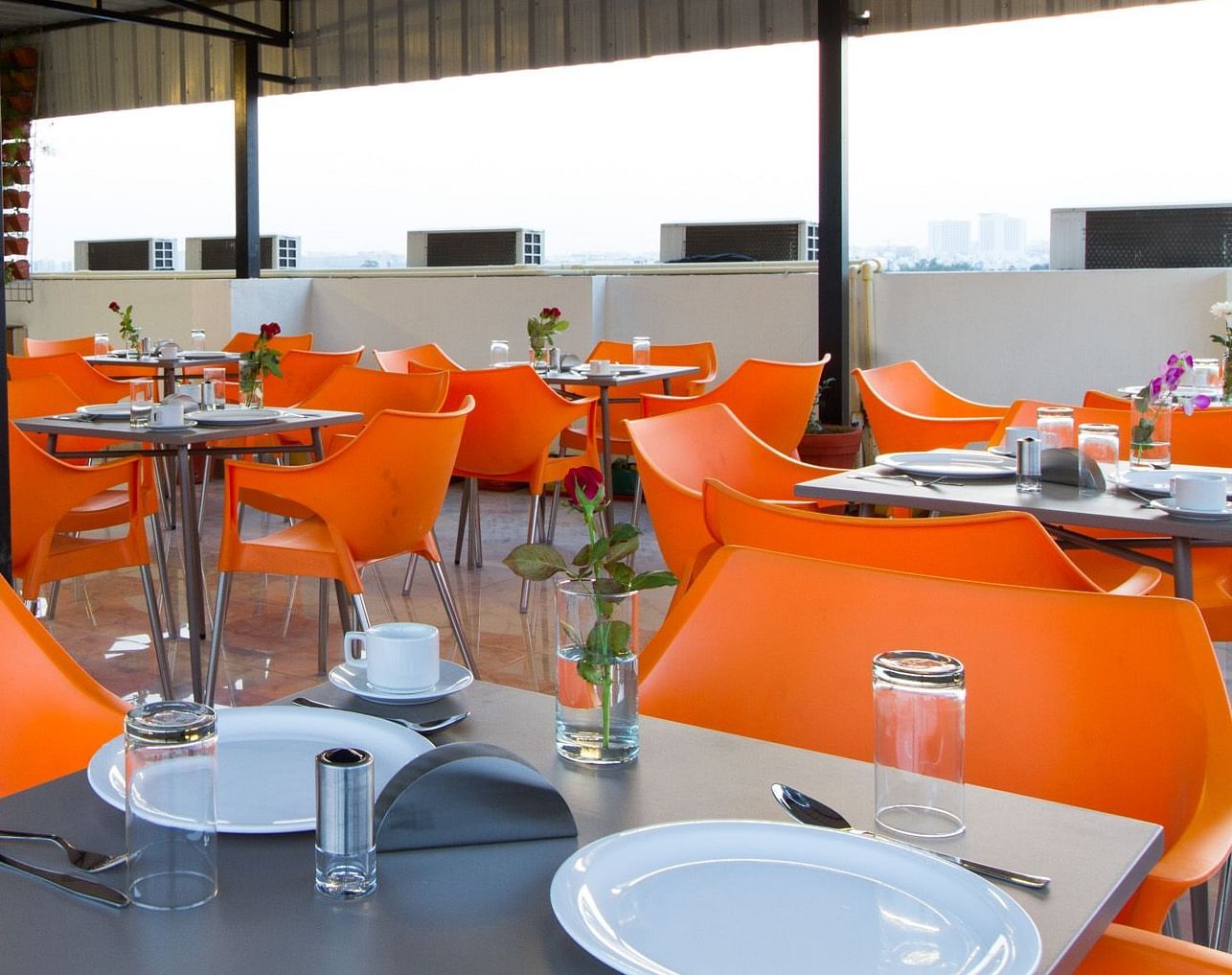 An outdoor dining space under a canopy offers numerous tables with bright orange seating at Sanctum Suites Whitefield, Bangalore.