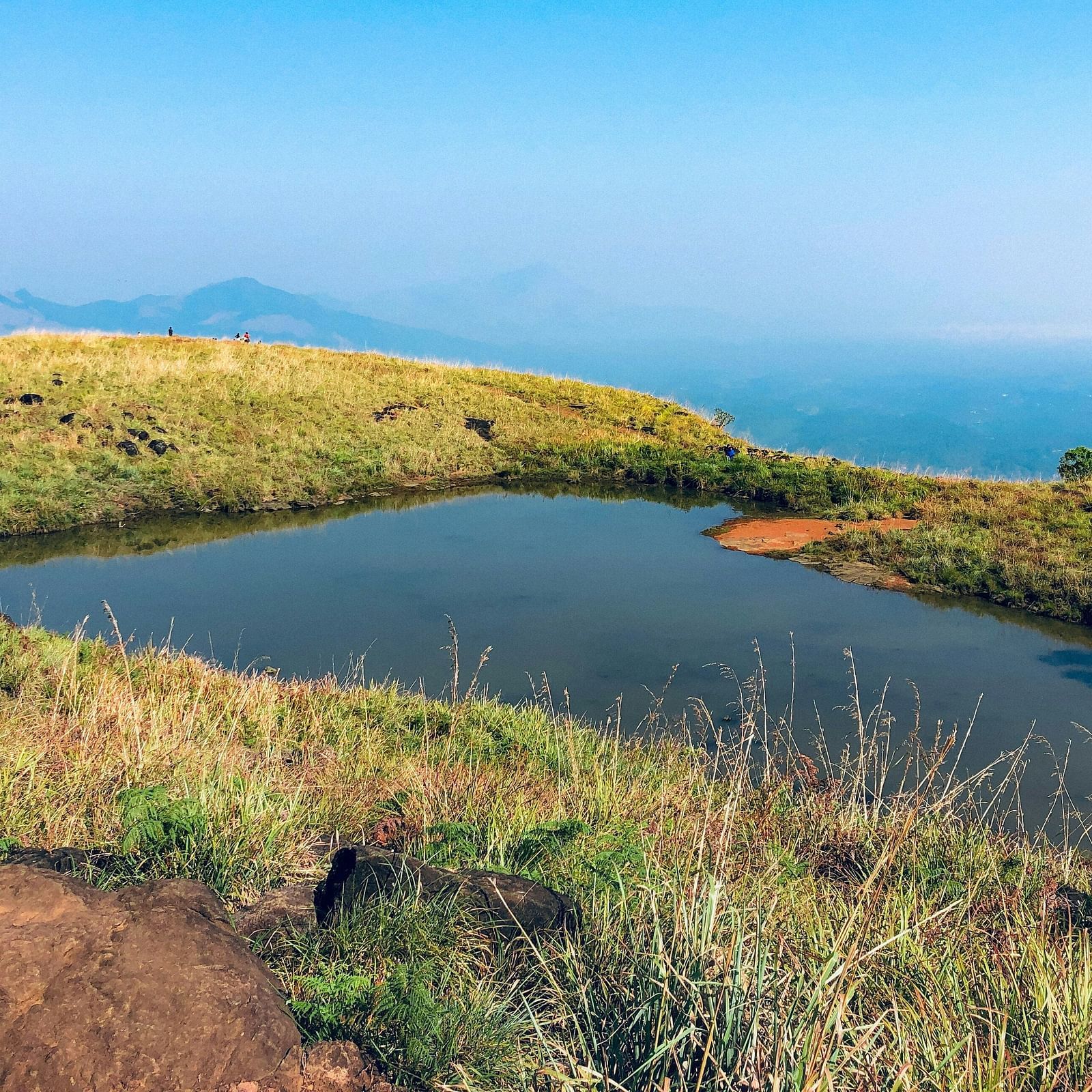 A high-altitude mountain landscape featuring a small pond surrounded by grassy terrain with distant mist-covered mountains under a bright blue sky.