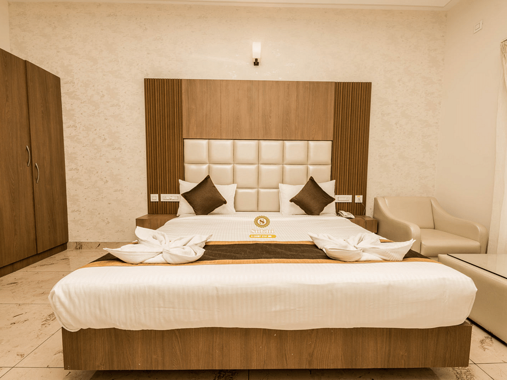Non AC Room featuring a king size bed with a cushioned headboard, two pillows, and a white duvet, with a wooden accent wall and furniture.- Sukhi - A Luxury Stay Inn, Chikkamagaluru