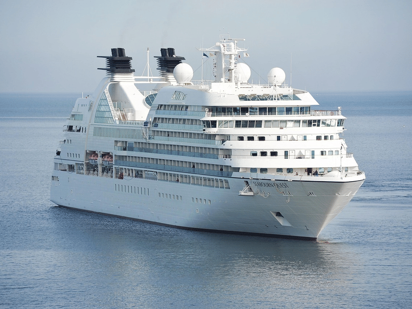 A large cruise ship sailing on calm waters, showing multiple decks, antennas, and lifeboats with the horizon visible in the distance.