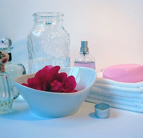 An image featuring a clear glass jar filled with rose petals sits in a bowl, surrounded by a stack of  towels, a perfume bottle, a spray bottle, and a bar of soap. 