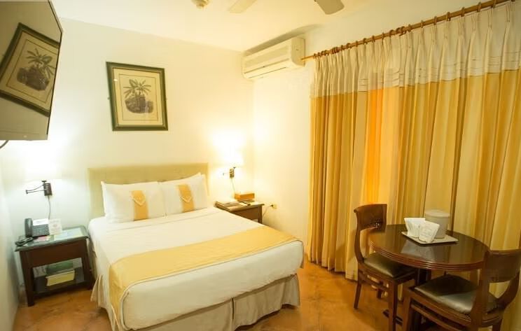 A bright bedroom with a bed, nightstands, and a dining table set near a window with yellow curtains | Economy Room | Altamont Court, Jamaica