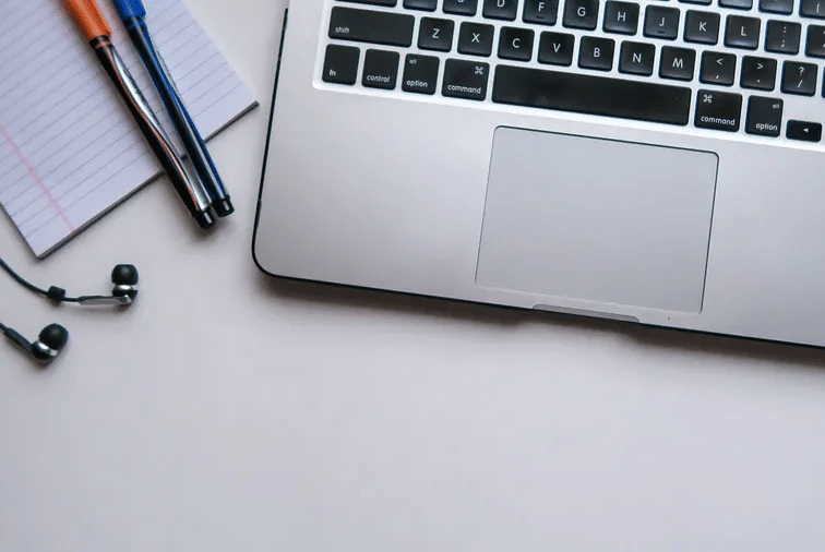 A laptop on a white desk beside a notepad, pens, and a pair of earphones neatly placed.