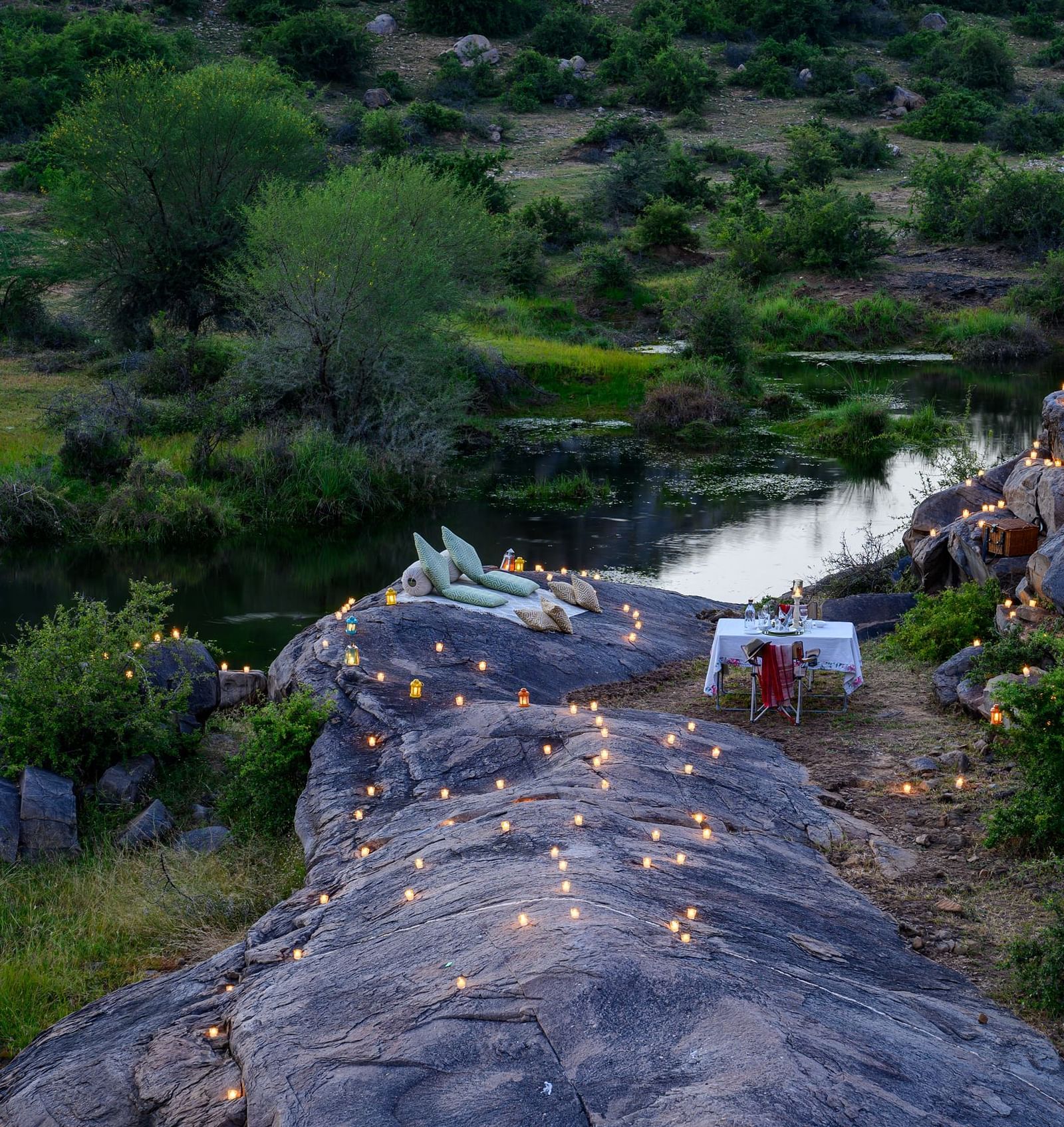 An image of an outdoor dining arrangement near a lake over viewing the mountains with bright lighting and greenery around - Utsav Camp Sariska