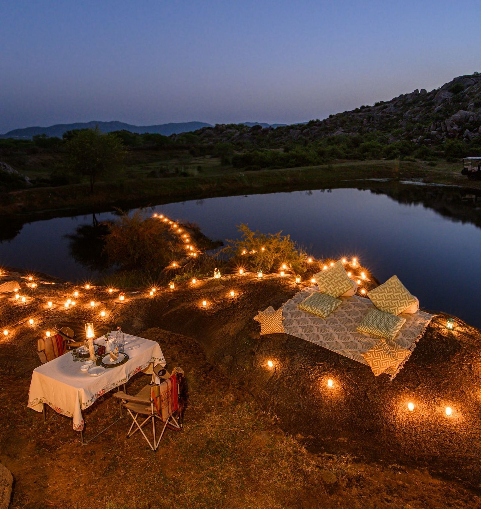 An image of an outdoor dining arrangement near a lake over viewing the mountains with bright lighting and greenery around - Utsav Camp Sariska