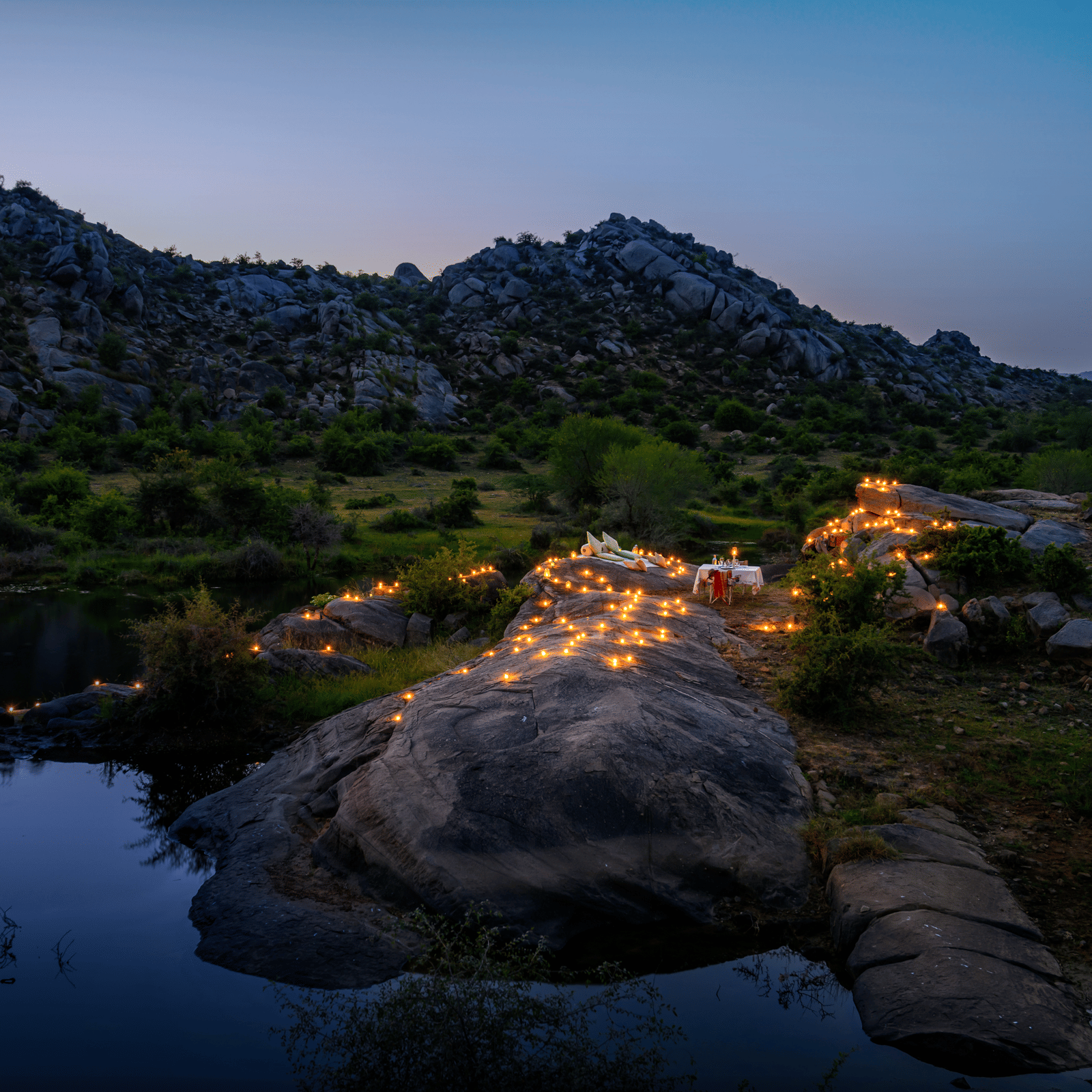 An image of an outdoor dining arrangement near a lake over viewing the mountains with bright lighting and greenery around - Utsav Camp Sariska