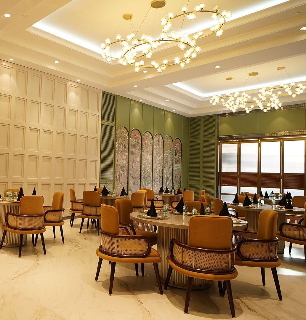  A stylish dining area at Nirvana Luxury Hotel, Ludhiana, featuring round tables with tan chairs, modern chandeliers, and elegant wall decor.