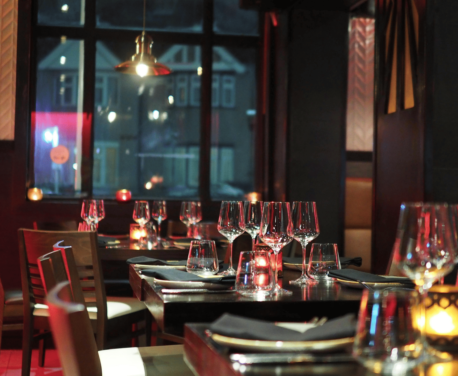 A moody interior shot of a restaurant dining table set with wine glasses and candles, seen through a dark window.