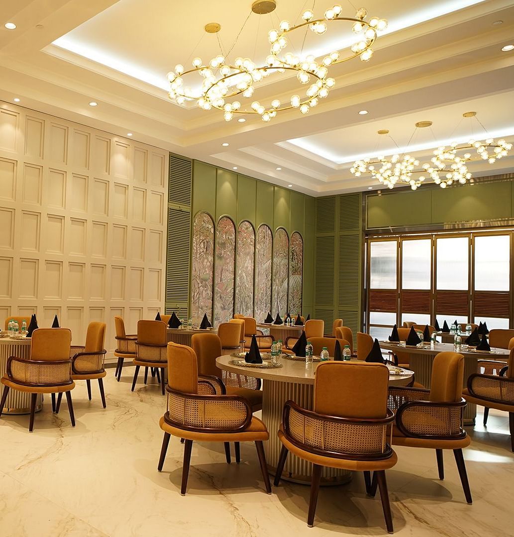  A stylish dining area at Nirvana Luxury Hotel, Ludhiana, featuring round tables with tan chairs, modern chandeliers, and elegant wall decor.