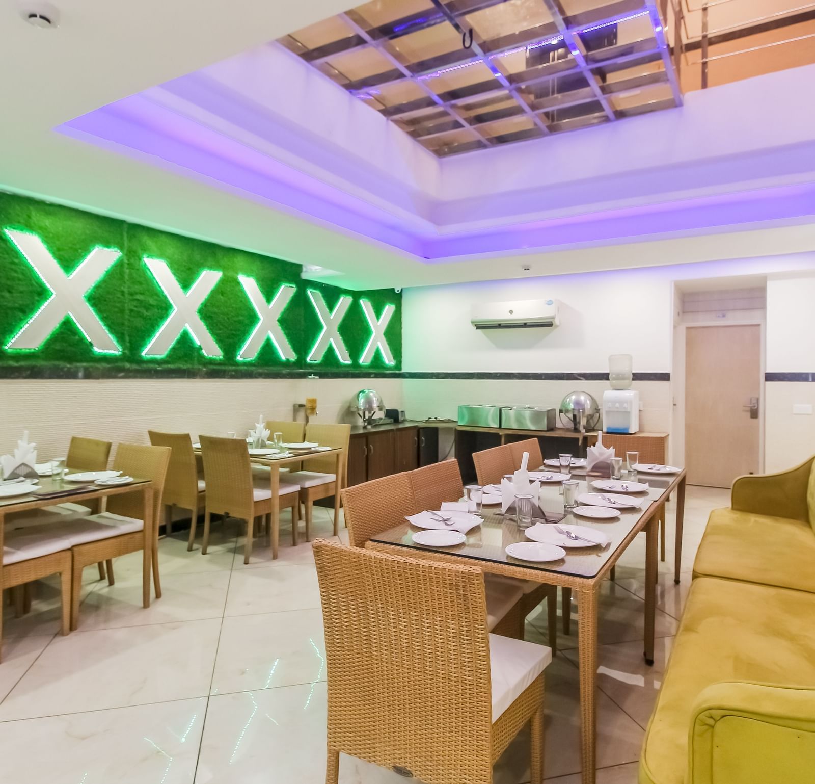 A restaurant interior with a long tufted sofa, wooden tables and chairs, a decorative wall design, and a tiled floor, lit by soft lighting. - Hotel Diamond Plaza, Chandigarh.