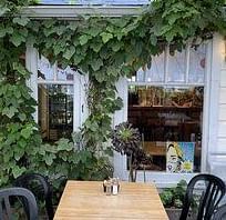 Exterior of a building with vines growing on the front, covering the windows and doorway. A table and chairs are visible on the sidewalk.