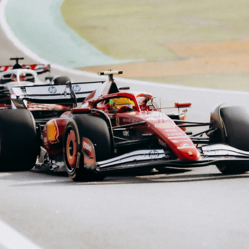 A view of the Ferrari and Haas Formula 1 cars on a track with the apex and green grass in view in the background.