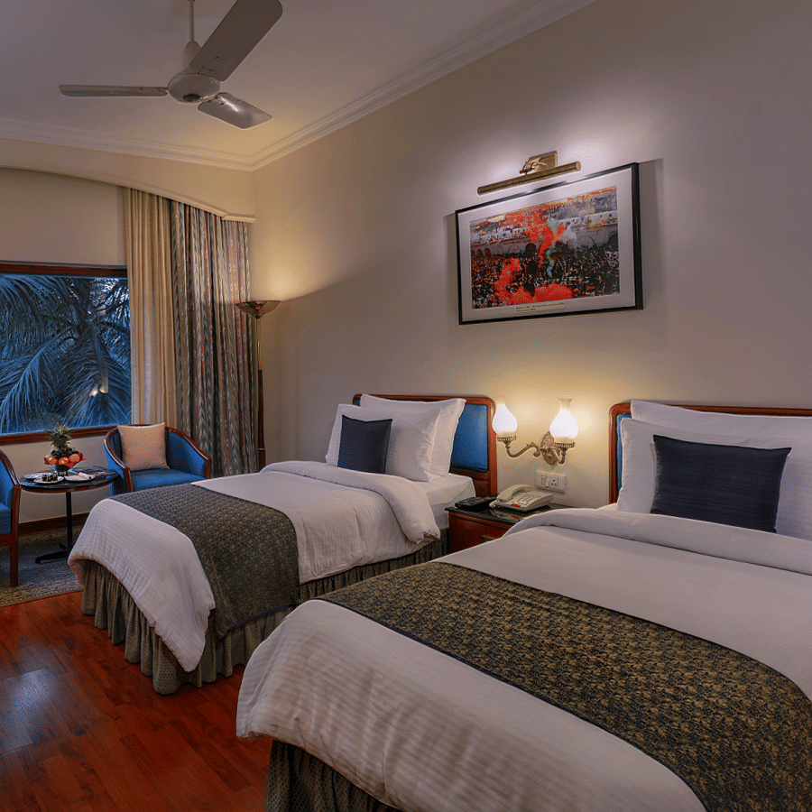 A Premiere Room at The Clarks Varanasi featuring twin beds, a ceiling fan, a wall-mounted television, and a seating area at The Clarks Varanasi.