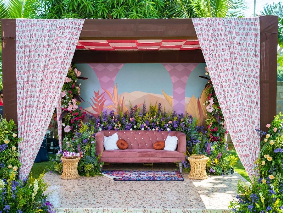 An outdoor wedding stage or mandap with a pink and white floral and fabric theme, featuring a seating area, set on a green lawn | Grande Pavillion | Grand Bay Resort and Spa