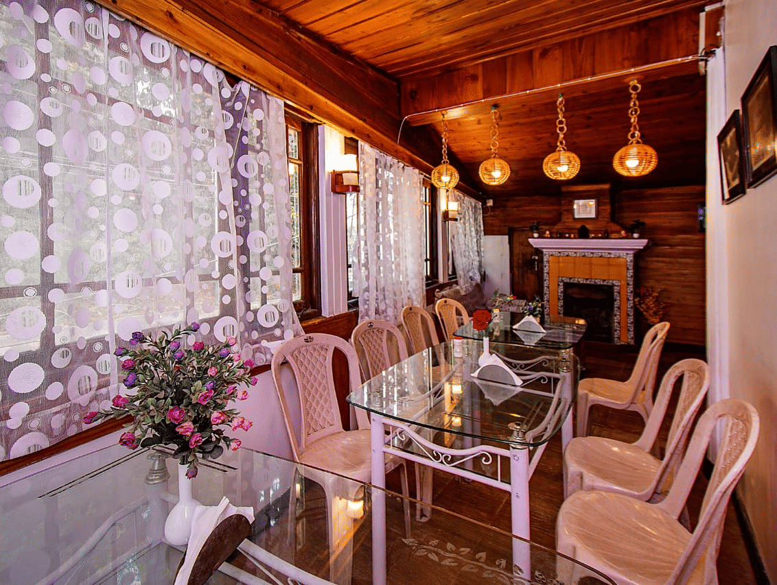 Dining area with a wooden ceiling, hanging lights, a glass table, and floral patterned white lace curtains at Hotel Dolphin Darjeeling.