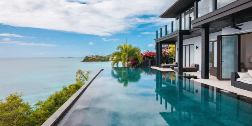 A modern multi-story villa with an expansive infinity pool stretching towards the stunning blue Caribbean sea and a distant island.