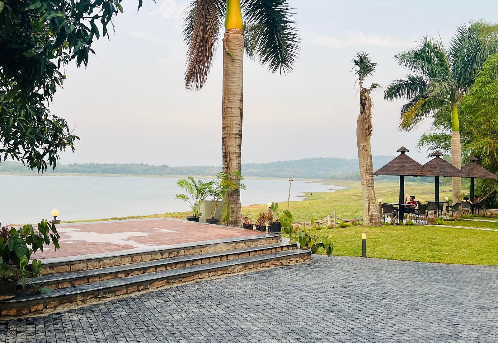 View of a lakeside area with stone steps, palm trees, and a paved pathway. A gazebo with a thatched roof sits near the water, with grassy areas and distant hills visible across the lake