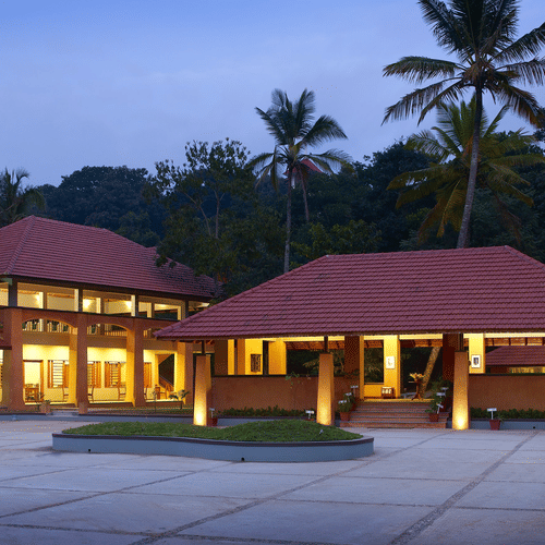 The facade of Abad Green Forest, Thekkady, featuring a single-storey structure with red sloping roofs, wide entrances, and a spacious open courtyard, set against a backdrop of lush greenery and tall palm trees.