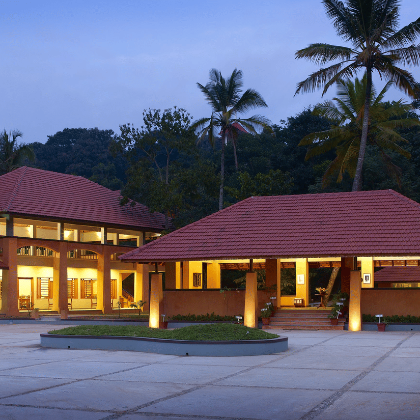 Facade view of the entrance to Abad Green Forest, Thekkady - A Forest Resort in Thekkady, during twilight hour with the lights on. An open area with a small patch of manicured garden in between and trees in the background area also in view.