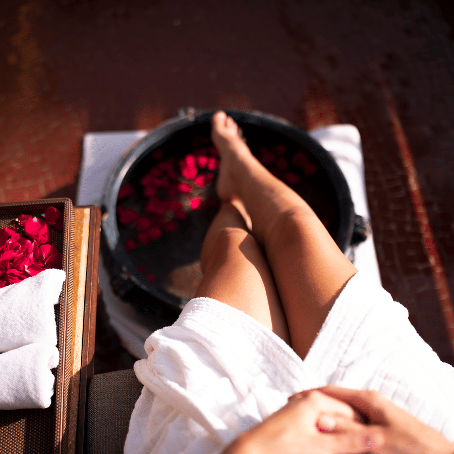 An image of a woman's leg resting in a round tub filled with water and rose petals, with towels neatly arranged on a tray beside it.